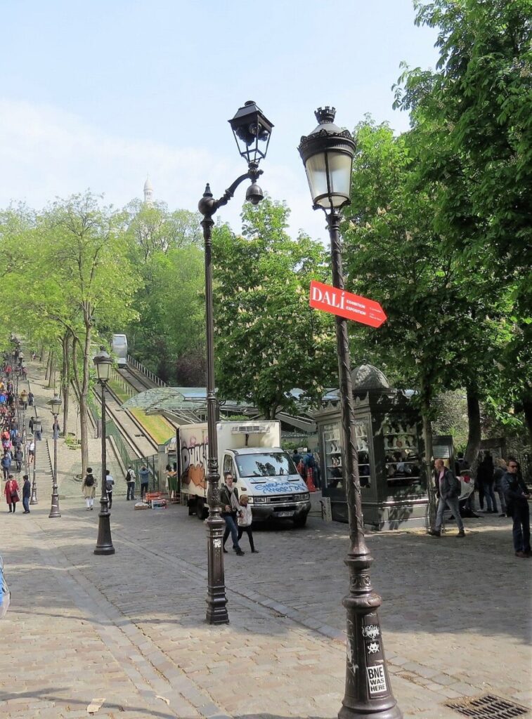 A ground-level view of the Montmartre Funicular's docking station, with vintage street lamps and a red 'DALÍ' sign, suggesting the cultural richness of the area.